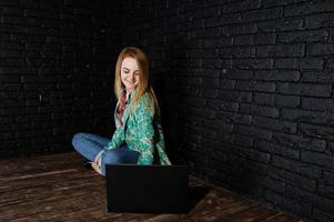 Stilvolles blondes Mädchen in Jacke und Jeans mit Laptop vor schwarzer Ziegelwand im Studio. foto