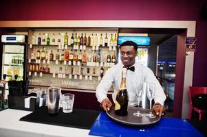 Afroamerikanischer Barkeeper an der Bar mit Champagner und Gläsern auf dem Tablett. foto