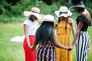 Gruppe von vier wunderschönen afroamerikanischen Frauen tragen Sommerhut, halten sich an den Händen und beten auf grünem Gras im Park. foto