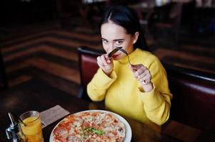 lustiges brünettes mädchen im gelben pullover, der pizza im restaurant isst. foto