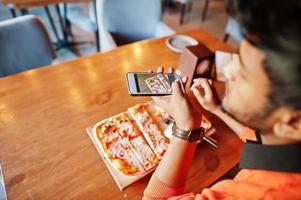 Selbstbewusster junger Inder in orangefarbenem Pullover, der in einer Pizzeria sitzt, Pizza isst und ein Foto auf seinem Handy macht.