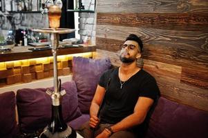 stylischer bart arabischer mann in brille und schwarzem t-shirt rauchen shisha indoor bar. Arabisches Model, das sich ausruht. foto