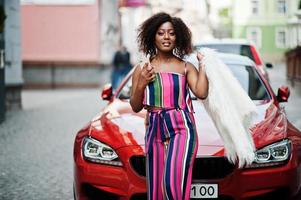 Modische Afroamerikanerin in rosa gestreiftem Overall mit flauschigem Kunstpelzmantel posierte mit Handy zur Hand gegen sattes rotes Luxusauto. foto