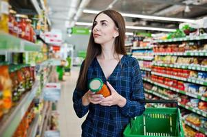 Einkaufsfrau, die die Regale im Supermarkt betrachtet. Porträt eines jungen Mädchens in einem Marktladen mit grünem Einkaufskorb und einer Dose Gemüse.