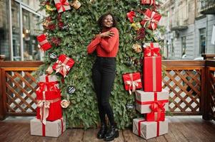 porträt einer lockigen afrikanerin mit modischem rotem rollkragenpullover, die sich gegen weihnachtsdekorationen posiert, silvesterthema. foto