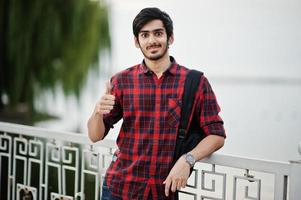 junger indischer student mann in kariertem hemd und jeans mit rucksack posierte auf abendstadt gegen see, zeigt daumen hoch. foto
