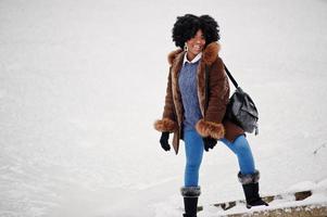 lockiges haar afroamerikanerin trägt schaffellmantel und handschuhe mit rucksack, der am wintertag gestellt wird. foto