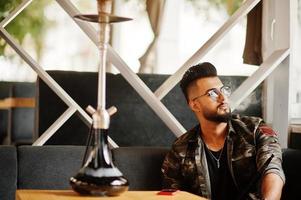 stylischer bart arabischer mann in brille und militärjacke raucht shisha an der straßenbar. Arabisches Model, das sich ausruht. foto