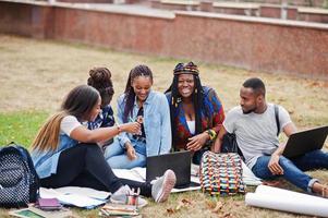 Gruppe von fünf afrikanischen College-Studenten, die gemeinsam Zeit auf dem Campus des Universitätshofs verbringen. schwarze Afro-Freunde sitzen auf Gras und lernen mit Laptops. foto