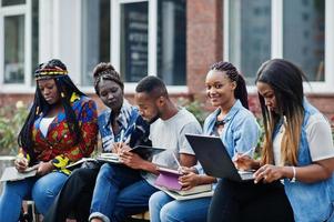 Gruppe von fünf afrikanischen College-Studenten, die gemeinsam Zeit auf dem Campus des Universitätshofs verbringen. schwarze afrofreunde, die an der bank mit schulsachen, laptops notizbüchern studieren. foto