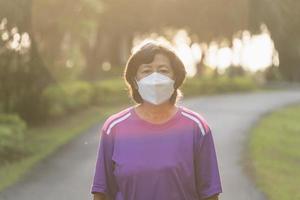 sportliche asiatische Seniorin der 60er Jahre mit OP-Maske und Joggen. Schöne ältere Asiatin, die an einem sonnigen Tag im Park läuft. foto