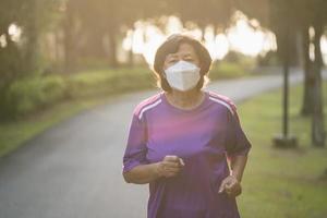 sportliche asiatische Seniorin der 60er Jahre mit OP-Maske und Joggen. Schöne ältere Asiatin, die an einem sonnigen Tag im Park läuft. foto