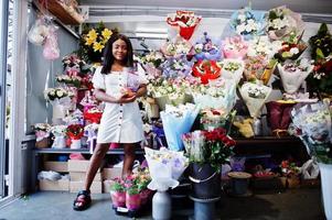 Schönes afrikanisch-amerikanisches Mädchen in zartem weißem Kleid mit Blumenstrauß in den Händen, die vor floralem Hintergrund im Blumenladen stehen. schwarze weibliche Floristin. foto