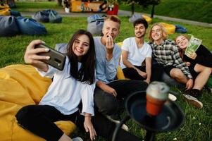 junge multiethnische gruppe von menschen, die filme im poof im open-air-kino ansehen und selfie am telefon machen. foto