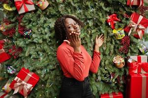 porträt einer lockigen afrikanerin mit modischem rotem rollkragenpullover, die sich gegen weihnachtsdekorationen posiert, silvesterthema. Luftkuss senden. foto