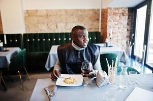 afrikanischer mann in schwarzer traditioneller kleidung sitzt im restaurant und isst pasta und trinkt wein. foto