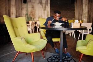 Modischer afroamerikanischer Mann in Anzug und Brille, der im Café sitzt und Salat isst. foto