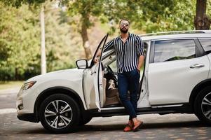 erfolgreicher arabischer mann in gestreiftem hemd und sonnenbrille posiert in der nähe seines weißen suv-autos. stilvolle arabische männer im transport. foto