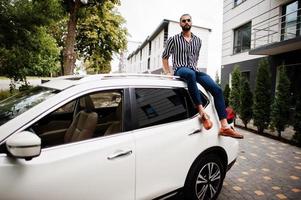 erfolgreicher arabischer mann trägt gestreiftes hemd und sonnenbrille, die auf dem dach seines weißen suv-autos sitzen. foto