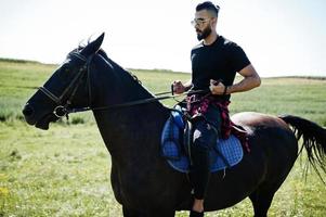 arabischer Mann mit hohem Bart in Schwarz und Sonnenbrille reitet auf einem arabischen Pferd. foto