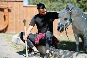 Arabischer Hipster-Bartmann-Holzfäller hält Axt gegen Ponypferd. foto