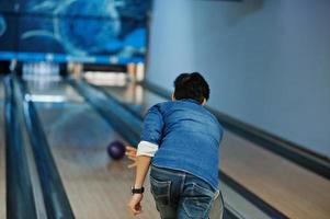 Rückseite des asiatischen Mannes im Jeanshemd, der mit Ball auf den Händen an der Bowlingbahn steht und ihn wirft. foto