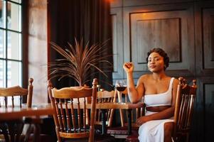 Afroamerikanerin, Retro-Frisur im weißen Kleid im Restaurant mit einem Glas Wein. foto