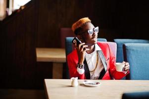 Mode-Afroamerikaner-Mann-Modell im roten Anzug, mit Highlights, Haaren und Sonnenbrillen, die im Café mit Kaffee sitzen und am Handy sprechen.