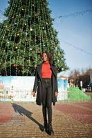 Porträt einer lockigen Afrikanerin mit modischem schwarzem Mantel und rotem Rollkragenpullover, die im Freien gegen den Hauptbaum des neuen Jahres der Stadt posiert. foto