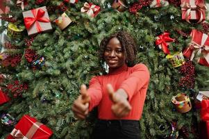 porträt einer lockigen afrikanerin mit modischem rotem rollkragenpullover, die sich gegen weihnachtsdekorationen posiert, silvesterthema. Daumen hoch zeigen. foto