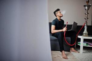 Schöner und modischer indischer Mann in Schwarz, der im Zimmer sitzt, Wasserpfeife raucht und am Laptop arbeitet. foto