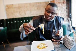 afrikanischer mann in schwarzer traditioneller kleidung sitzt im restaurant und isst pasta und trinkt wein. foto