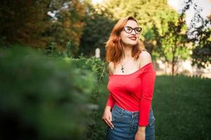 attraktive rothaarige frau mit brille, tragen auf roter bluse und jeansrock, die im grünen park posieren. foto