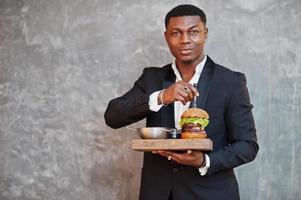 respektabler junger afroamerikaner im schwarzen anzug hält tablett mit doppelburger gegen graue wand. foto