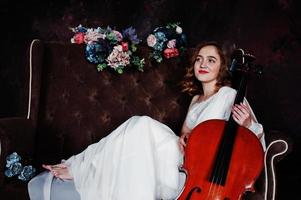 hübscher junger Mädchenmusiker im weißen Kleid mit Kontrabass, der auf braunem Vintage-Sofa sitzt. foto