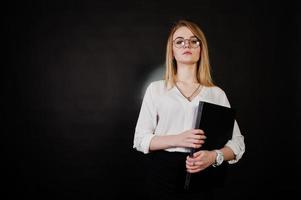 Studioporträt einer blonden Geschäftsfrau mit Brille, weißer Bluse und schwarzem Rock, die einen Laptop vor dunklem Hintergrund hält. erfolgreiche frau und stilvolles mädchenkonzept. foto