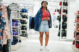 Modische afroamerikanische Frau in rotem Hemd und Jeansrock mit Jacke posierte im Bekleidungsgeschäft. Es ist Zeit zum Einkaufen. foto