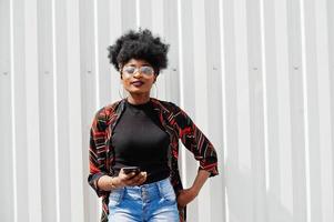 afrikanische frau mit afrohaar, in jeansshorts und brille, die mit handy in der hand gegen eine weiße stahlwand posiert. foto