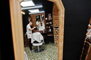 junger bärtiger mann, der vom friseur die haare schneiden lässt, während er im friseursalon auf dem stuhl sitzt. Barbier Seele. foto