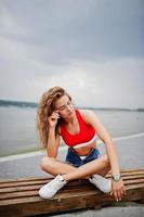 sexy lockiges Model-Mädchen in rotem Oberteil, Jeans-Denim-Shorts, Brille und Turnschuhen posiert auf einer Bank am See. foto