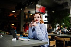 Fröhliche junge schöne rothaarige Frau mit Brille, die ihr Telefon, Touchpad und Notebook benutzt, während sie an ihrem Arbeitsplatz im Café mit einer Tasse Kaffee sitzt. foto