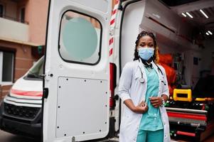 Afroamerikanische Sanitäterin mit medizinischer Gesichtsschutzmaske, die vor einem Krankenwagen steht. foto
