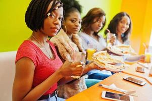 Vier junge afrikanische Mädchen in einem bunten Restaurant mit Pizzastücken auf Teller und Säften. foto