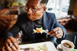 Modischer afroamerikanischer Mann in Anzug und Brille, der im Café sitzt und Salat isst. foto