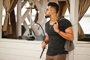 stylischer bart arabischer mann in brille und schwarzem t-shirt rauchen wasserpfeife im freien. Arabisches Model, das sich ausruht. foto