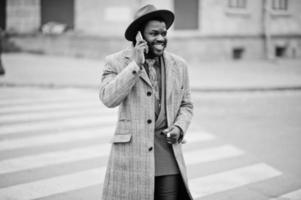 Stilvolles afroamerikanisches Mannmodell in grauem Mantel, Jackenkrawatte und rotem Hut, das auf dem Zebrastreifen geht und mit dem Handy spricht. Schwarz-Weiß-Foto. foto