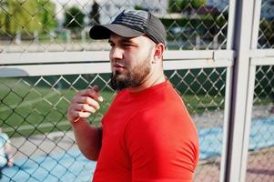 junger brutaler bärtiger muskulöser mann trägt rotes hemd, shorts und mütze im stadion. foto