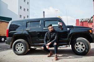 modereicher bart arabischer mann trägt schwarze jeansjacke und sonnenbrille, die gegen großes schwarzes suv-auto gestellt werden. stylischer, erfolgreicher und modischer arabischer Modeltyp. foto