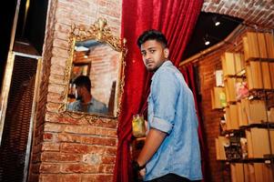 Porträt eines gutaussehenden, erfolgreichen, bärtigen, südasiatischen, jungen indischen Freiberuflers in blauem Jeanshemd, der im Café mit Cocktail vor dem Spiegel steht. foto