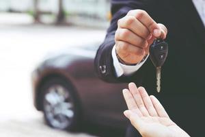 Geschäftsmann Hand hält Autoschlüssel vorne mit Neuwagen im Hintergrund. Parkplatz vor dem Haus. Transportkonzept. Lassen Sie Platz zum Kopieren, um Nachrichtentext zu schreiben. foto